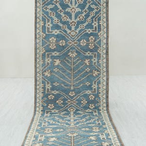 May include: A rectangular, blue runner rug with a detailed, symmetrical pattern. The rug features a central design of stylized trees and floral motifs in shades of beige and brown. The border is a darker brown with a cream-colored edge.