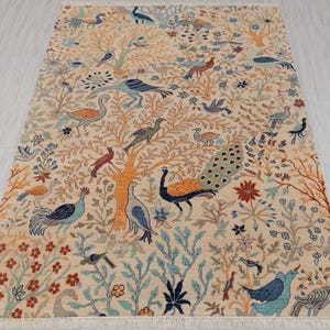 以下が含まれることがあります： クリーム色の敷物で、孔雀、鳥、木が細かく描かれた模様が施されています。敷物はフリンジの縁取りがされています。