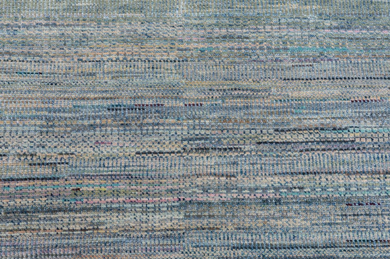 May include: Close-up of a textured rug with a mix of blue, gray, and beige hues. The rug's design features horizontal and vertical lines, creating a woven appearance. The color palette includes hints of teal and pink.