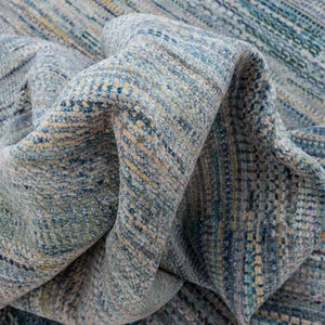 May include: A close-up of a folded rug with a textured, woven design. The rug features a blend of blue, beige, and yellow hues, creating a subtle, striped pattern. The fabric appears soft and pliable, with visible details in the weave.