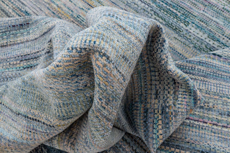 May include: A close-up of a folded rug with a textured, woven design. The rug features a blend of blue, beige, and yellow hues, creating a subtle, striped pattern. The fabric appears soft and pliable, with visible details in the weave.