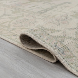 May include: Close-up of a cream-colored rug with a subtle, light green and pink floral pattern. The rug is rolled up, showing the edge and the soft texture of the fabric.