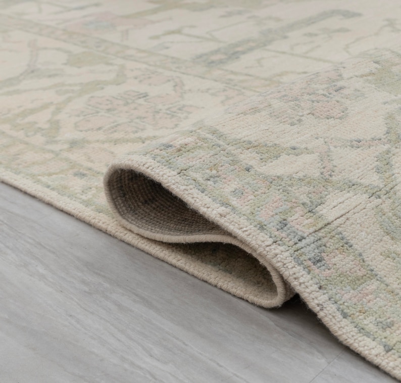 May include: Close-up of a cream-colored rug with a subtle, light green and pink floral pattern. The rug is rolled up, showing the edge and the soft texture of the fabric.