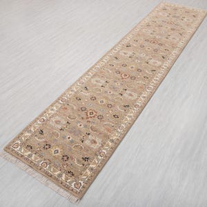 Op de afbeelding: Een rechthoekige beige loper met een ingewikkeld bloemenpatroon. De loper heeft een crèmekleurige rand en franjes aan beide uiteinden. Het ontwerp bevat verschillende kleuren zoals bruin, rood en blauw.
