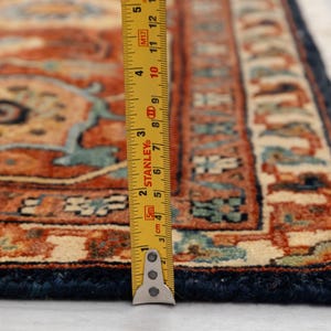 May include: Close-up of a section of an intricately patterned rug. The rug features a complex design in shades of rust, beige, and teal, with a dark blue border. A yellow measuring tape is placed on the rug, showing inches.