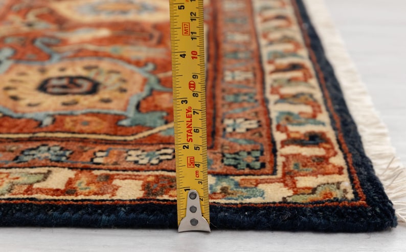 May include: Close-up of a section of an intricately patterned rug. The rug features a complex design in shades of rust, beige, and teal, with a dark blue border. A yellow measuring tape is placed on the rug, showing inches.