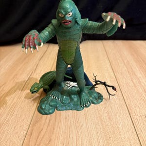May include: A green Creature from the Black Lagoon figurine with red accents on its hands and mouth. The figurine stands on a green base with a lizard, a skeleton hand, and the text "THE CREATURE".