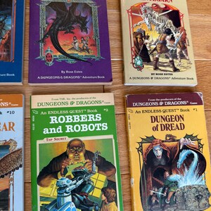 May include: A collection of vintage "Dungeons & Dragons" adventure books. The covers feature fantasy illustrations of dragons, robots, and dungeons. Titles include "Dragon of Doom," "Pillars of Pentegarn," "Robbers and Robots," and "Dungeon of Dread."