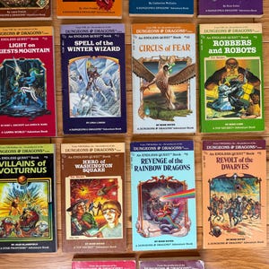 May include: A collection of vintage "Dungeons & Dragons" adventure books. The books feature colorful covers with fantasy illustrations, including dragons, wizards, and other mythical creatures. Titles include "Light on Quests Mountain" and "Revolt of the Dwarves."