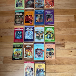 May include: A collection of vintage "Dungeons & Dragons" adventure books, featuring colorful covers with fantasy illustrations. Titles include "Return to Brookmere," "Dragon's Wing," and "Revolt of the Dwarves." The books are arranged on a wooden surface.