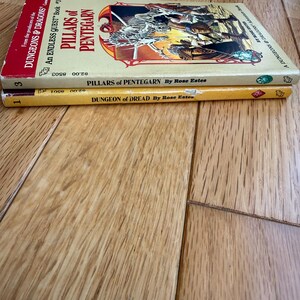 May include: Two vintage paperback books stacked on a wooden surface. The top book is titled "Pillars of Pentegarn" and the bottom book is "Dungeon of Dread". Both books are part of the "Dungeons & Dragons" series.