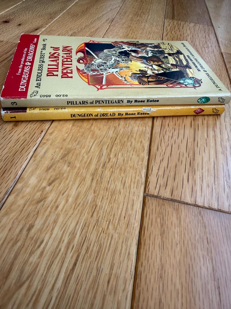 May include: Two vintage paperback books stacked on a wooden surface. The top book is titled "Pillars of Pentegarn" and the bottom book is "Dungeon of Dread". Both books are part of the "Dungeons & Dragons" series.