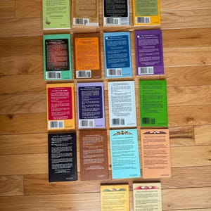 May include: A collection of paperback books with colorful covers, arranged on a wooden surface. The books feature various colors, including green, orange, blue, and purple. The backs of the books are visible, displaying text and bar codes. The books are part of the "Pick a Path to Adventure" series.