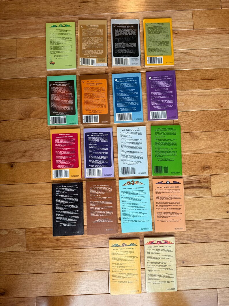 May include: A collection of paperback books with colorful covers, arranged on a wooden surface. The books feature various colors, including green, orange, blue, and purple. The backs of the books are visible, displaying text and bar codes. The books are part of the "Pick a Path to Adventure" series.
