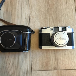 May include: A silver Aires 35mm film camera with a black leather case. The camera has a black leather strap and a black lens cap.