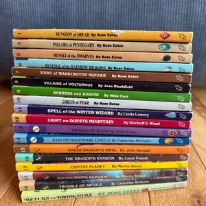 May include: A stack of vintage paperback books with colorful spines. Titles include "Dungeon of Dread," "Revolt of the Dwarves," and "Dragon of Doom." The books are arranged on a wooden surface, with a blue fabric background.