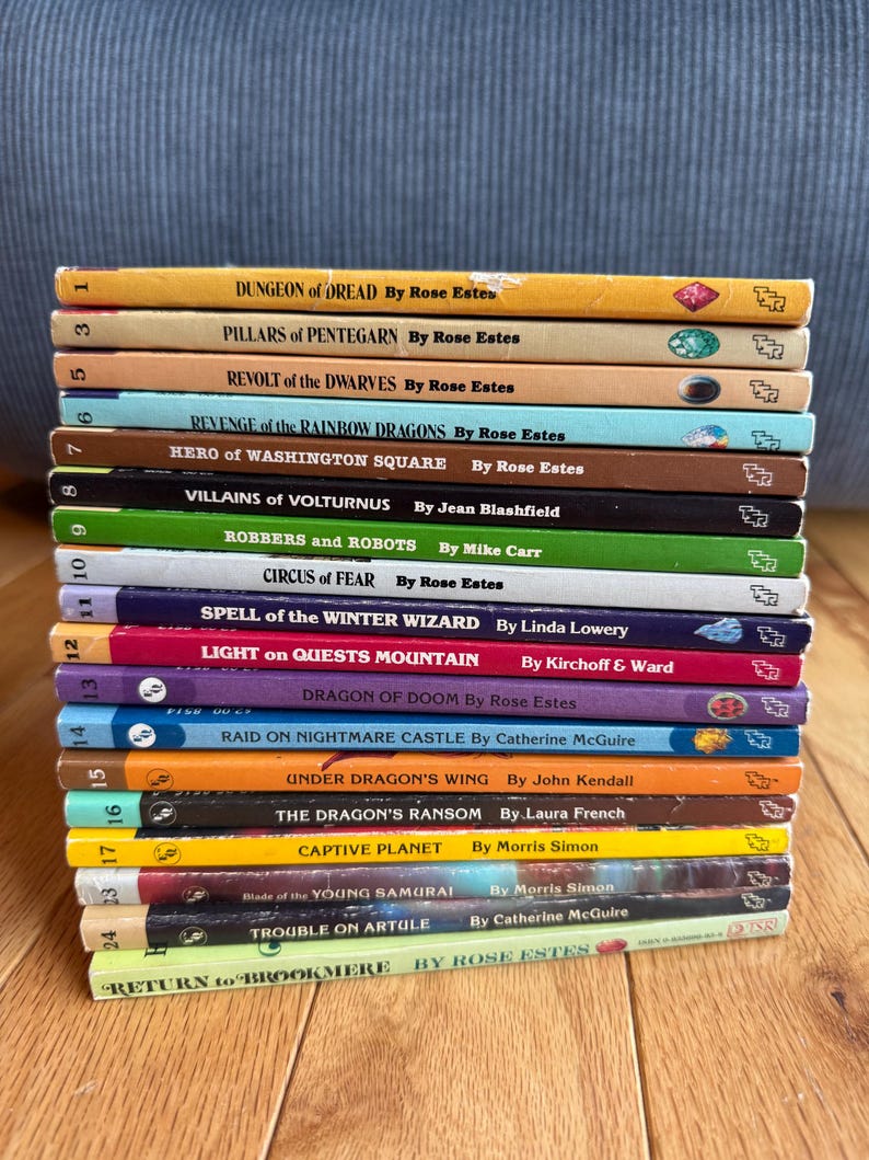 May include: A stack of vintage paperback books with colorful spines. Titles include "Dungeon of Dread," "Revolt of the Dwarves," and "Dragon of Doom." The books are arranged on a wooden surface, with a blue fabric background.