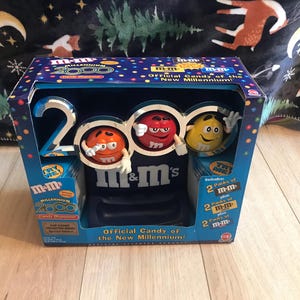 May include: A blue and red box with a picture of three M&M's characters on it. The box says "Official Candy of the New Millennium!" and "m&m's Millennium 2000".
