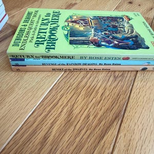 May include: A stack of three paperback books with colorful covers. The top book is titled "Return to Brookmere" with a fantasy illustration. Below are "Revenge of the Rainbow Dragons" and "Revolt of the Dwarves" by Rose Estes. The books are on a wooden surface.