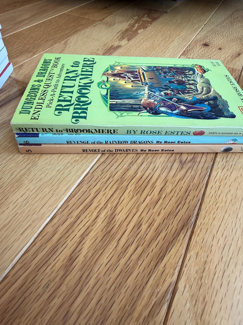 May include: A stack of three paperback books with colorful covers. The top book is titled "Return to Brookmere" with a fantasy illustration. Below are "Revenge of the Rainbow Dragons" and "Revolt of the Dwarves" by Rose Estes. The books are on a wooden surface.