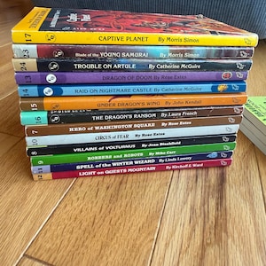 May include: A stack of vintage paperback books with colorful covers. Titles include "Captive Planet," "Blade of the Young Samurai," and "Trouble on Artule." The books are arranged on a wooden surface, showcasing their spines and titles.