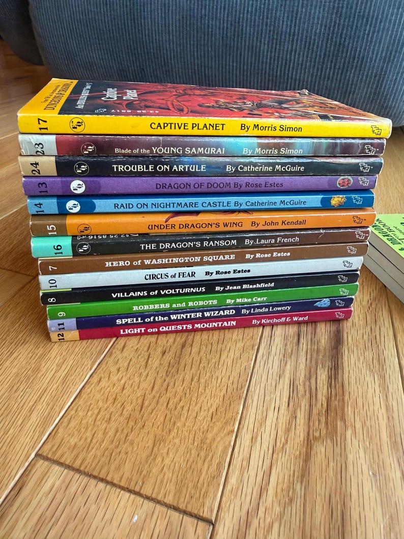 May include: A stack of vintage paperback books with colorful covers. Titles include "Captive Planet," "Blade of the Young Samurai," and "Trouble on Artule." The books are arranged on a wooden surface, showcasing their spines and titles.