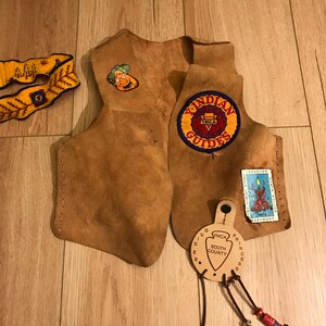 May include: A brown leather vest with a "YMCA Indian Guides" patch, a "South County" patch, and a "Induction Ceremony" patch. The vest has a beaded strap with a leather tassel.