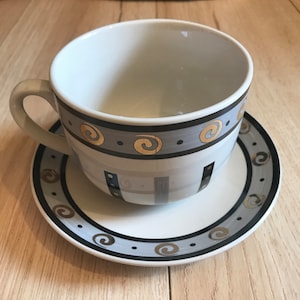 May include: A white ceramic teacup and saucer set with a gray and black geometric pattern and gold accents. The cup has a handle and the saucer has a scalloped edge.