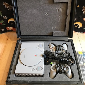 May include: A gray Sony PlayStation console with two gray controllers, a power cord, and an AV cable, all inside a black hard-shell carrying case.