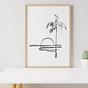 Puede incluir: Un dibujo en blanco y negro de una palmera con el sol poni&eacute;ndose detr&aacute;s. El dibujo est&aacute; enmarcado en un marco de madera claro y est&aacute; sobre una mesa blanca.