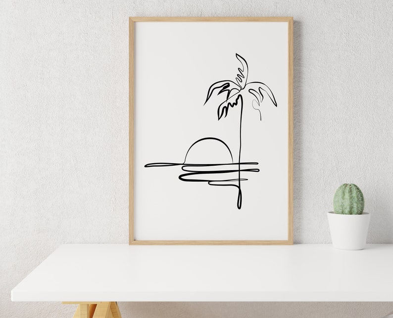 Puede incluir: Un dibujo en blanco y negro de una palmera con el sol poni&eacute;ndose detr&aacute;s. El dibujo est&aacute; enmarcado en un marco de madera claro y est&aacute; sobre una mesa blanca.