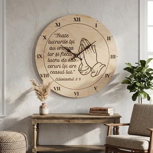 Op de afbeelding: Grote, ronde houten wandklok met Romeinse cijfers en zwarte wijzers. De klok heeft een illustratie van biddende handen en tekst in een scriptlettertype. De klok is aan een lichtgekleurde muur bevestigd.