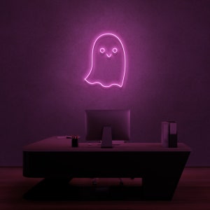 May include: A pink neon ghost sign with a smiling face, hanging on a purple wall above a black desk with a computer monitor and a stack of folders.