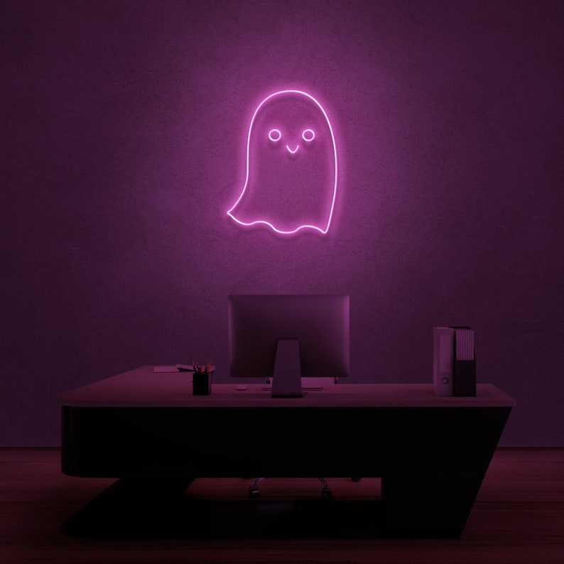 May include: A pink neon ghost sign with a smiling face, hanging on a purple wall above a black desk with a computer monitor and a stack of folders.