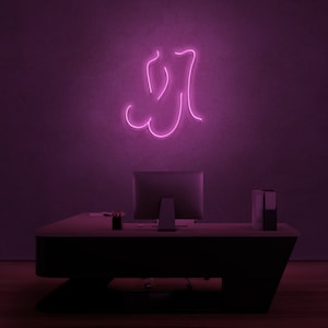 Puede incluir: Un letrero de neón rosa que representa un contorno estilizado de una persona sentada. El letrero está montado en una pared morada sobre un escritorio negro con un monitor de computadora y una pila de carpetas.