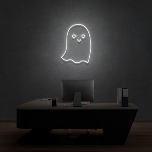 May include: A white neon sign shaped like a ghost with a smiling face, hanging on a gray wall above a black desk with a computer monitor and a stack of folders.