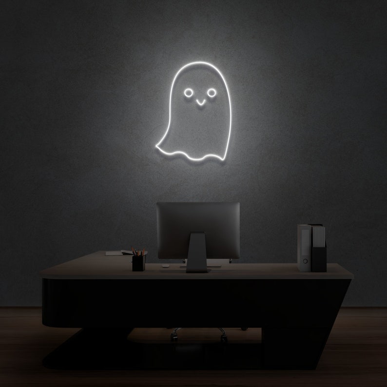 May include: A white neon sign shaped like a ghost with a smiling face, hanging on a gray wall above a black desk with a computer monitor and a stack of folders.