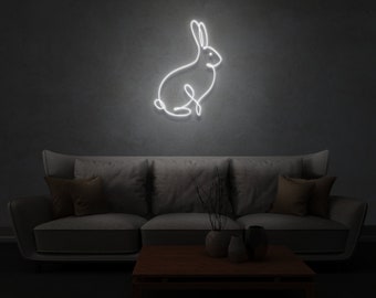 May include: White neon sign depicting a simple line drawing of a rabbit sitting on a dark gray wall. The sign is above a gray couch with brown throw pillows.
