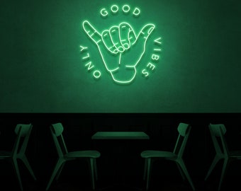 May include: Green neon sign with a hand gesture and the text "GOOD VIBES ONLY" in a circle. The sign is hanging on a green wall above a dark wooden table and chairs.