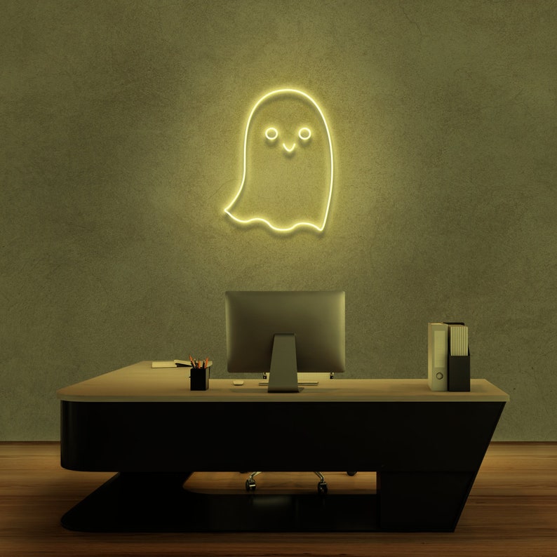 May include: A yellow neon ghost sign with a smiling face hangs on a green wall above a black desk with a computer monitor and a stack of folders.