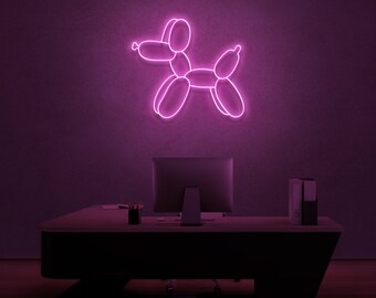 May include: Pink neon sign shaped like a balloon animal dog. The sign is mounted on a wall above a desk with a computer monitor and office supplies.
