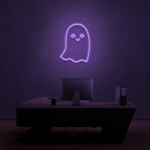May include: A purple neon sign shaped like a ghost with a smiling face. The sign is mounted on a wall above a desk with a computer monitor and a stack of folders.