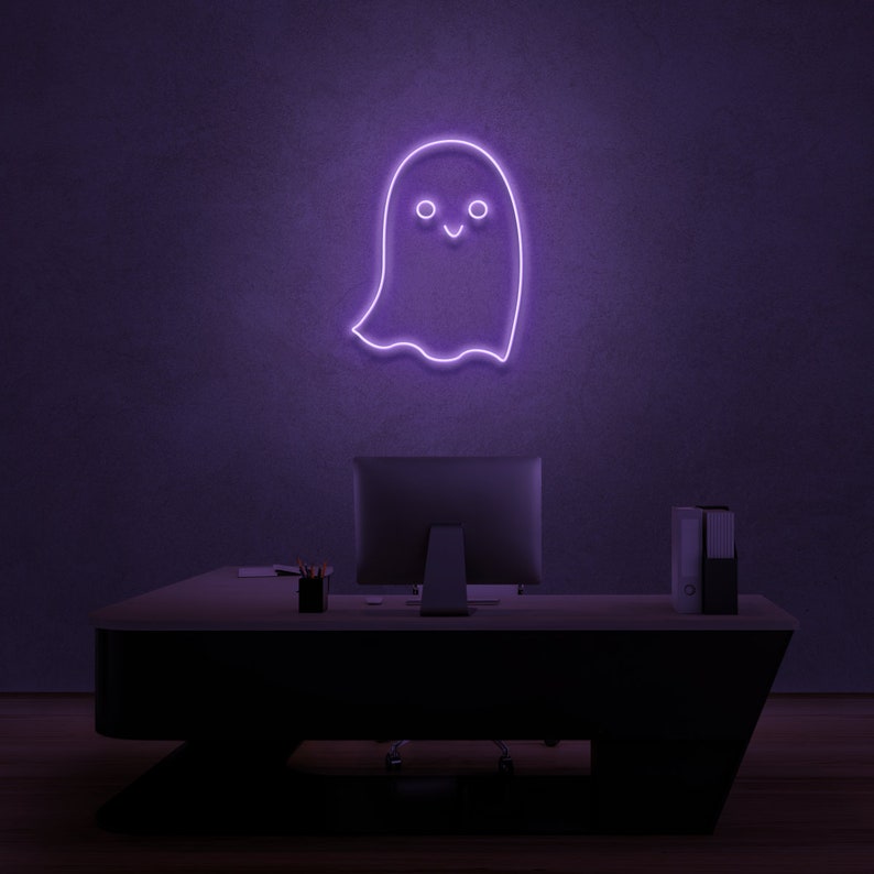 May include: A purple neon sign shaped like a ghost with a smiling face. The sign is mounted on a wall above a desk with a computer monitor and a stack of folders.