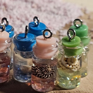 Può includere: Una collezione di otto piccole bottiglie di vetro con tappi di vari colori e cime argentate. Ogni bottiglia contiene una piccola conchiglia e  è riempita di un liquido trasparente. Le bottiglie sono disposte su una superficie di legno.