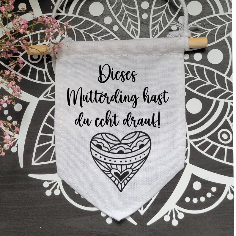 Op de afbeelding: Witte stoffen banner met zwarte tekst die "Dieses Mutterding hast du echt drauf!" luidt en een zwart-wit hartontwerp.