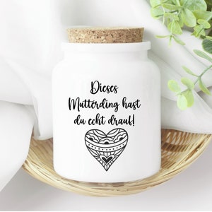 Könnte beinhalten: Weiße Keramikdose mit Korkverschluss und einem schwarz-weißen Herzdesign. Die Dose hat den Text "Dieses Mutterding hast du echt drauf!" aufgedruckt.