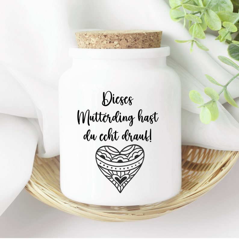 Op de afbeelding: Witte keramische pot met een kurken deksel en een zwart-wit hartontwerp. De pot heeft de tekst "Dieses Mutterding hast du echt drauf!" erop gedrukt.