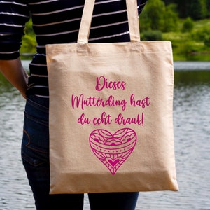Op de afbeelding: Een beige tote bag met een roze hartontwerp en de tekst "Dieses Mutterding hast du echt drauf!" erop gedrukt.