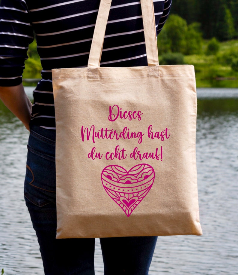 Op de afbeelding: Een beige tote bag met een roze hartontwerp en de tekst "Dieses Mutterding hast du echt drauf!" erop gedrukt.