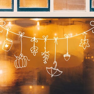 Op de afbeelding: Een witte string met herfstdecoraties hangt voor een raam. De decoraties omvatten een pompoen, een eikel, een pot met een esdoornblad, een tak met bladeren, een tak met bessen en een paraplu.
